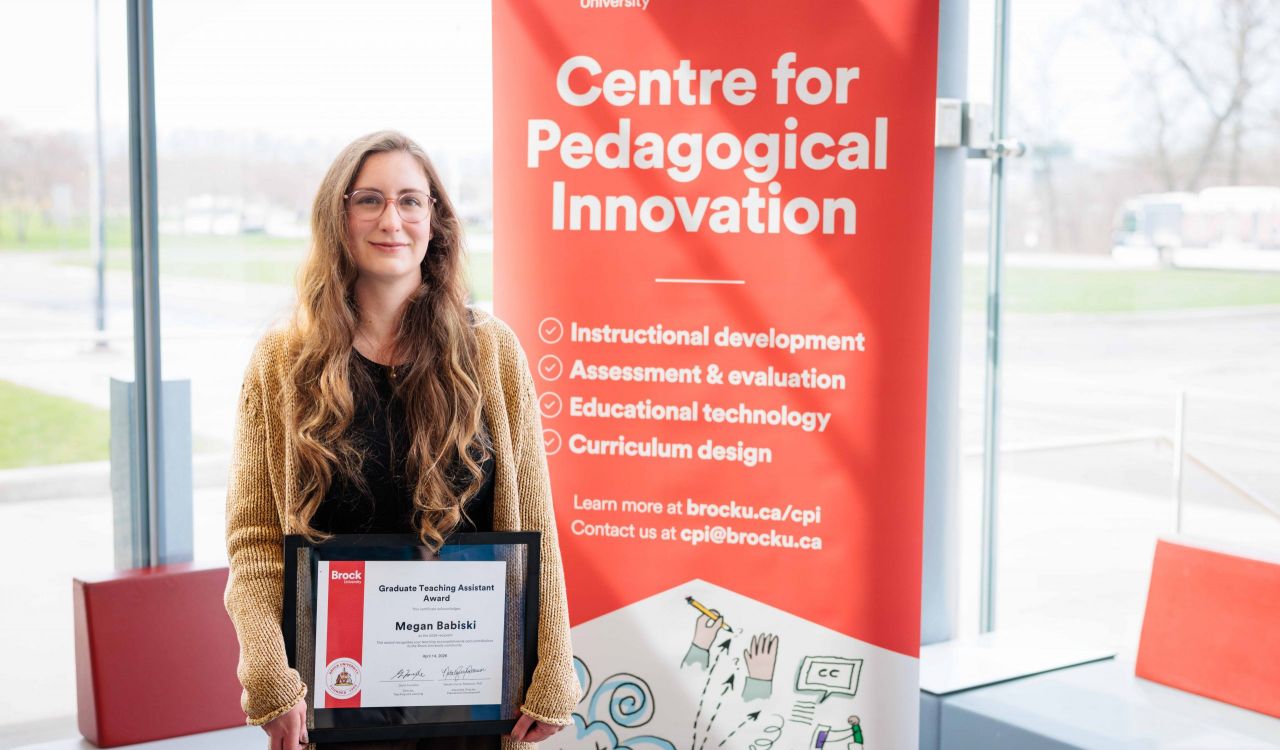 Megan Babiski holds her Graduate Teaching Assistant certificate next to a banner advertising Brock's Centre for Pedagogical Innovation.