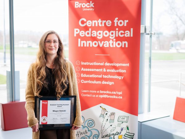 Megan Babiski holds her Graduate Teaching Assistant certificate next to a banner advertising Brock's Centre for Pedagogical Innovation.