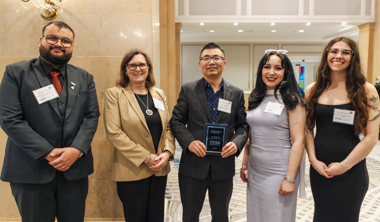 Five people standing in a line pose for a photo. The man in the centre is holding an award.