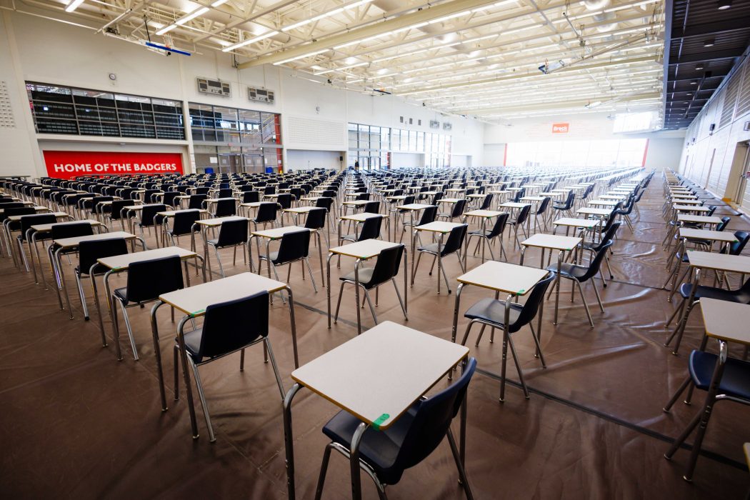 Wide view of a large indoor gymnasium set up for exams, with hundreds of individual desks and chairs spaced in long rows beneath bright overhead lights.
