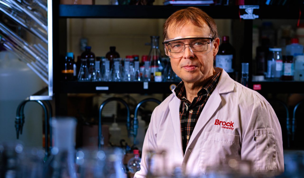 Close-up of Travis Dudding standing in his laboratory, with fuzzed-out rows of bottles in the foreground and background.