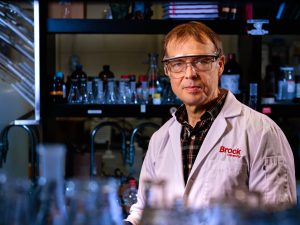 Close-up of Travis Dudding standing in his laboratory, with fuzzed-out rows of bottles in the foreground and background.