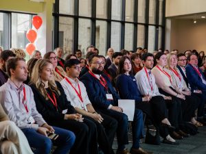 A large audience sits in an event space watching someone off camera speak.
