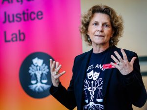 A speaker gestures while speaking beside a Health, Art, and Justice Lab banner.