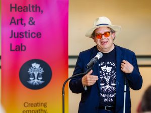 A speaker addresses the audience at a microphone beside a Health, Art and Justice Lab banner.
