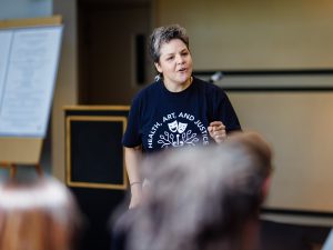 A speaker wearing a Health, Art, and Justice T-shirt addresses an audience during a presentation.