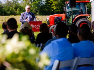 A speaker addresses an outdoor audience from a Brock University podium beside a red tractor during an event in a field.