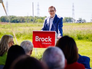 A smiling speaker stands at a Brock University podium while addressing an outdoor audience in a sunny field.