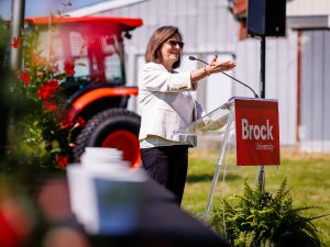A woman speaks at a Brock University podium outdoors, with a red tractor and event flowers visible behind her.