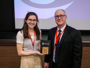 Two people stand together, one holding an award.