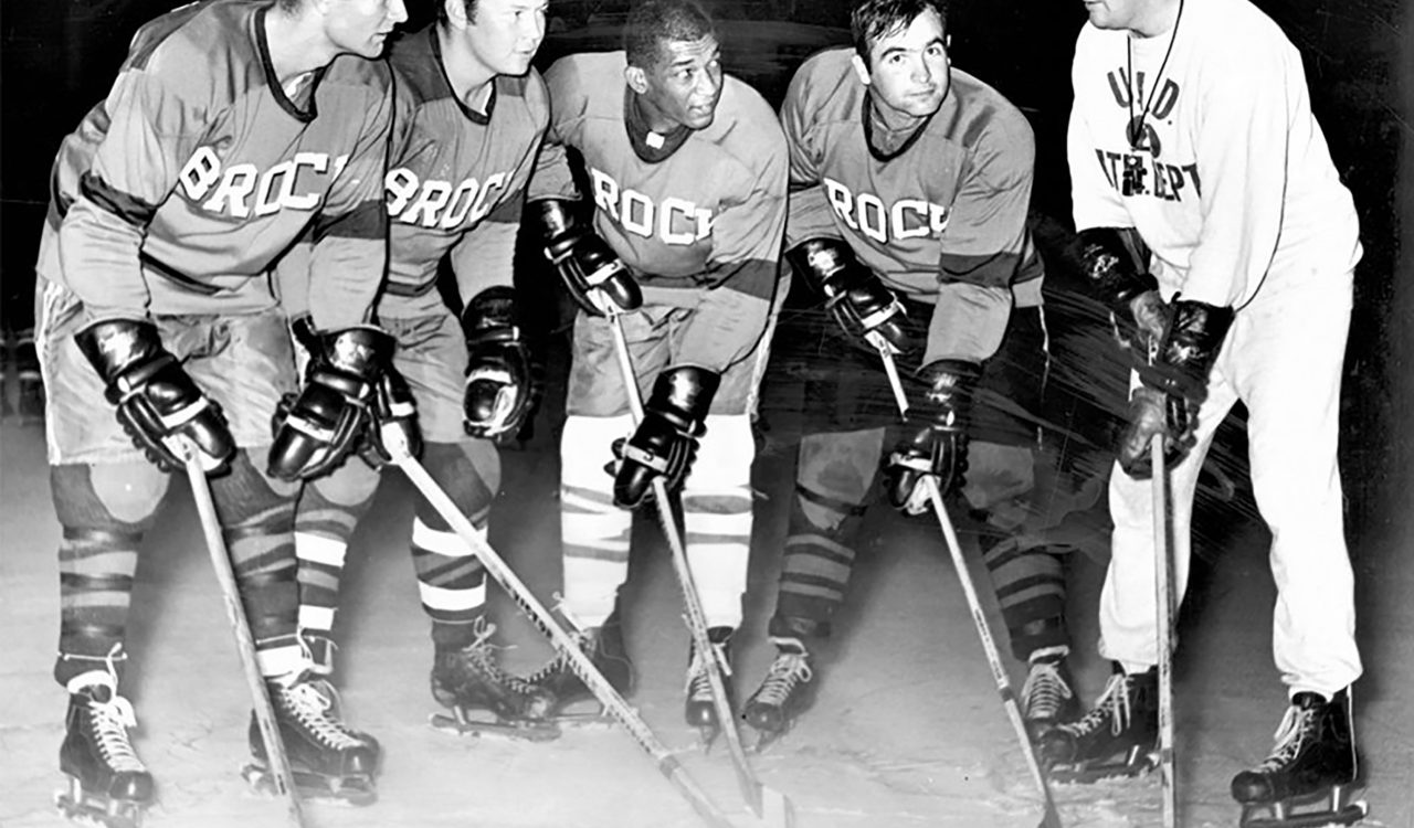 Four ice hockey players look towards their coach while placing their hockey sticks on the ice.