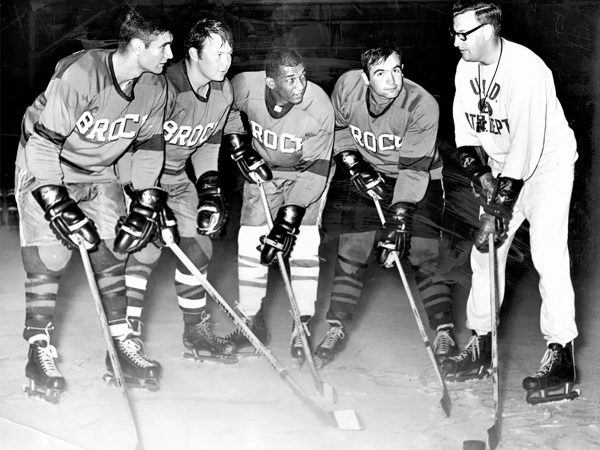 Four ice hockey players look towards their coach while placing their hockey sticks on the ice.
