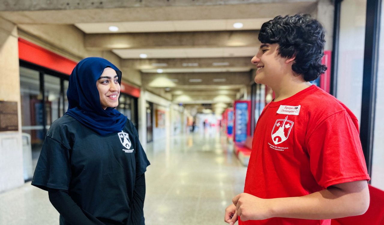 Two people stand together talking and smiling in a university hallway.