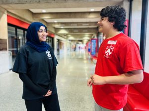 Two people stand together talking and smiling in a university hallway.