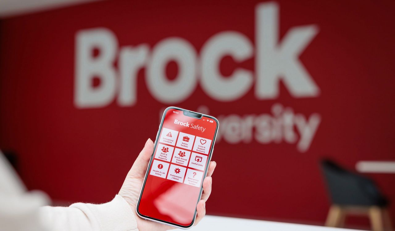 A hand holds a smartphone displaying the Brock Safety app, with a blurred red Brock University sign in the background.