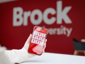 A hand holds a smartphone displaying the Brock Safety app, with a blurred red Brock University sign in the background.