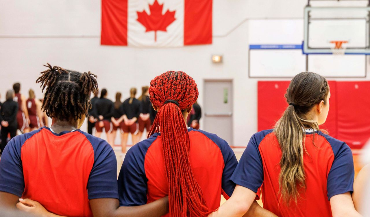 Three female athletes are seen from the back with their arms around each other's backs while standing in a gymnasium.