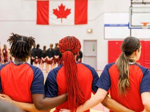 Three female athletes are seen from the back with their arms around each other's backs while standing in a gymnasium.