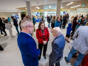 Three people have a conversation while other mingle around them at a community event.