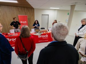 An audience listens to three people speaking at an information table during a community event.