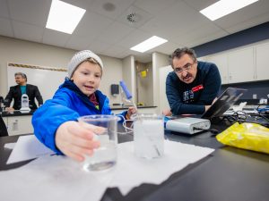 An educator watches while a child picks up a container of water in a classroom to do an experiment.