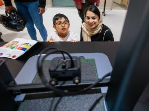 A mother and child watch a 3D printer work at a library event.