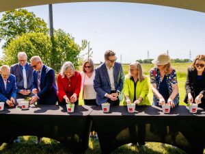 A large group of people stand beside a table under a tent on a sunny day with a field behind them.