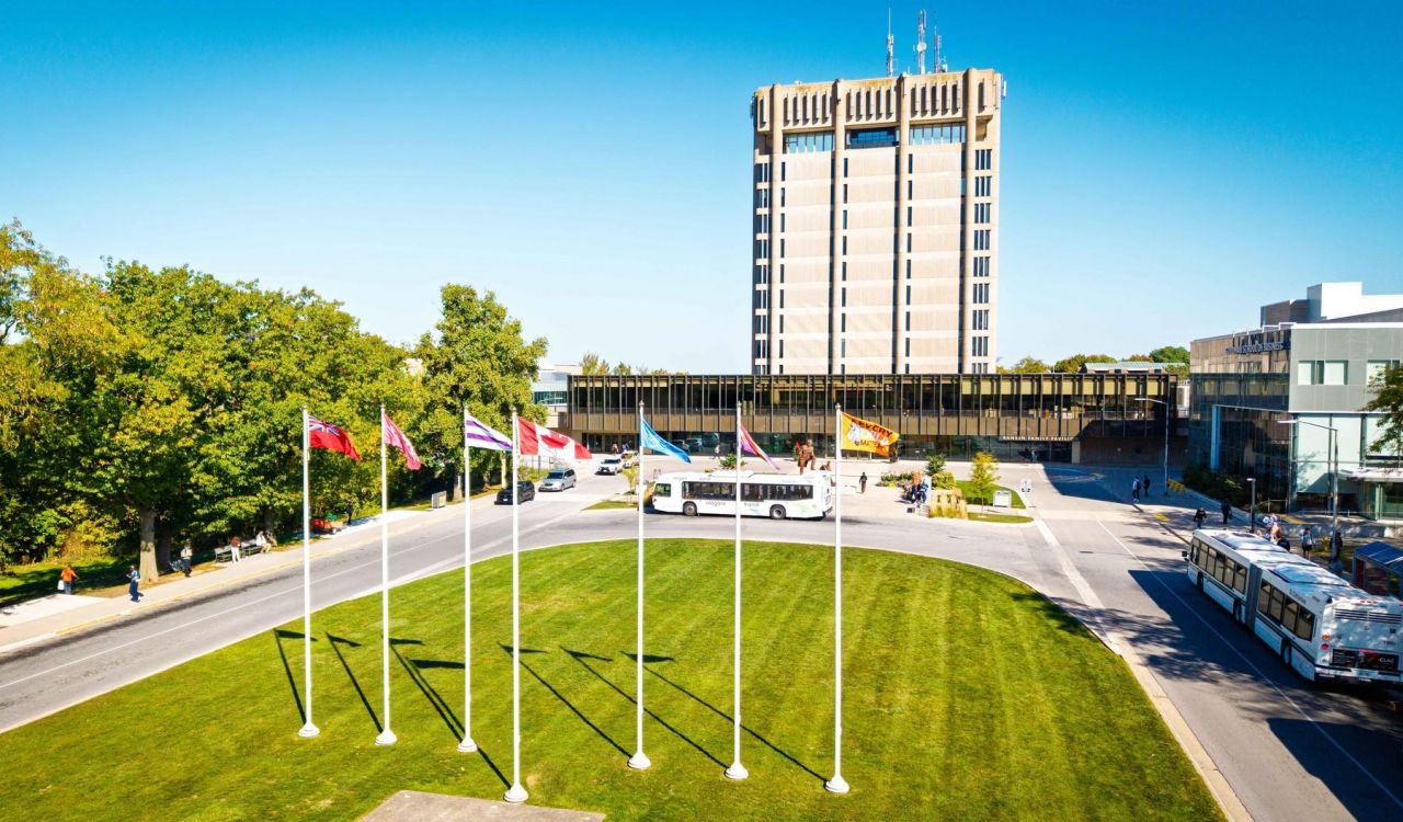 Wide view of the Rankin Family Pavillion and Schmon Tower at Brock University with a circular green lawn, five flagpoles, campus roads and buses on a clear day.
