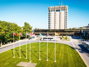 Wide view of the Rankin Family Pavillion and Schmon Tower at Brock University with a circular green lawn, five flagpoles, campus roads and buses on a clear day.
