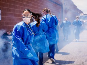 Nursing student in blue protective gown stands in smoke as other nursing students move through an outdoor Code Orange emergency simulation.