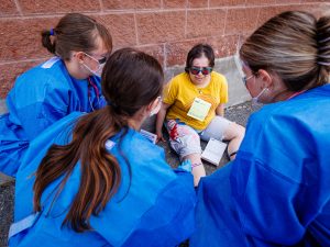 Nursing students in blue protective gowns assess a seated simulated patient with visible mock injuries during an emergency training scenario.