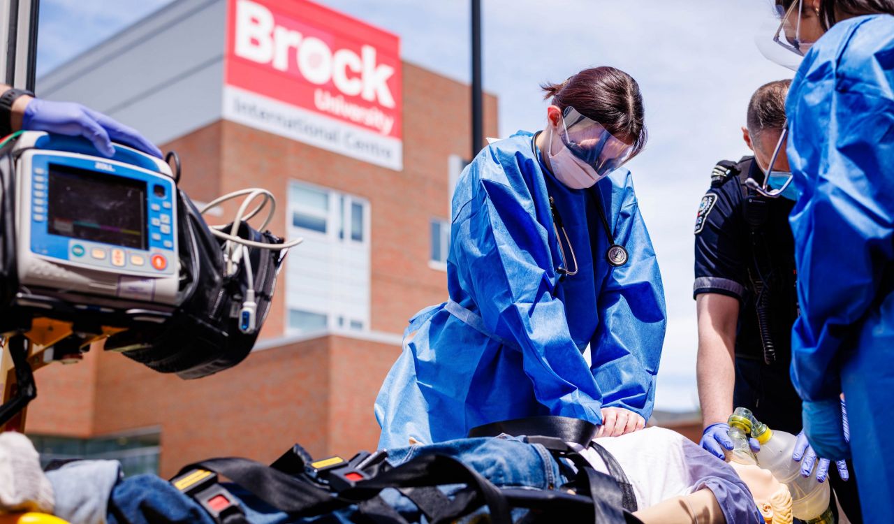 Nursing students in blue protective gowns provide simulated emergency care beside Niagara EMS paramedics outside Brock University’s International Centre.