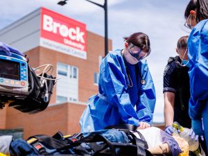 Nursing students in blue protective gowns provide simulated emergency care beside Niagara EMS paramedics outside Brock University’s International Centre.