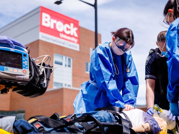Nursing students in blue protective gowns provide simulated emergency care beside Niagara EMS paramedics outside Brock University’s International Centre.