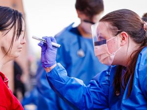 Nursing student in blue protective gear uses a penlight to assess a simulated patient’s eyes during an outdoor emergency training scenario.