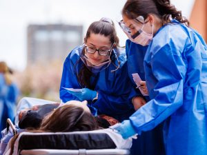 Two nursing students in blue protective gowns assess a simulated patient lying on a stretcher during emergency response training.