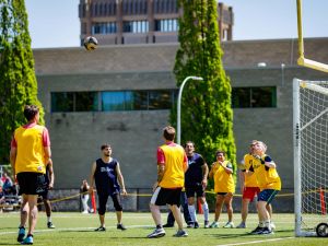 People play soccer on a field at a university with buildings in the background.