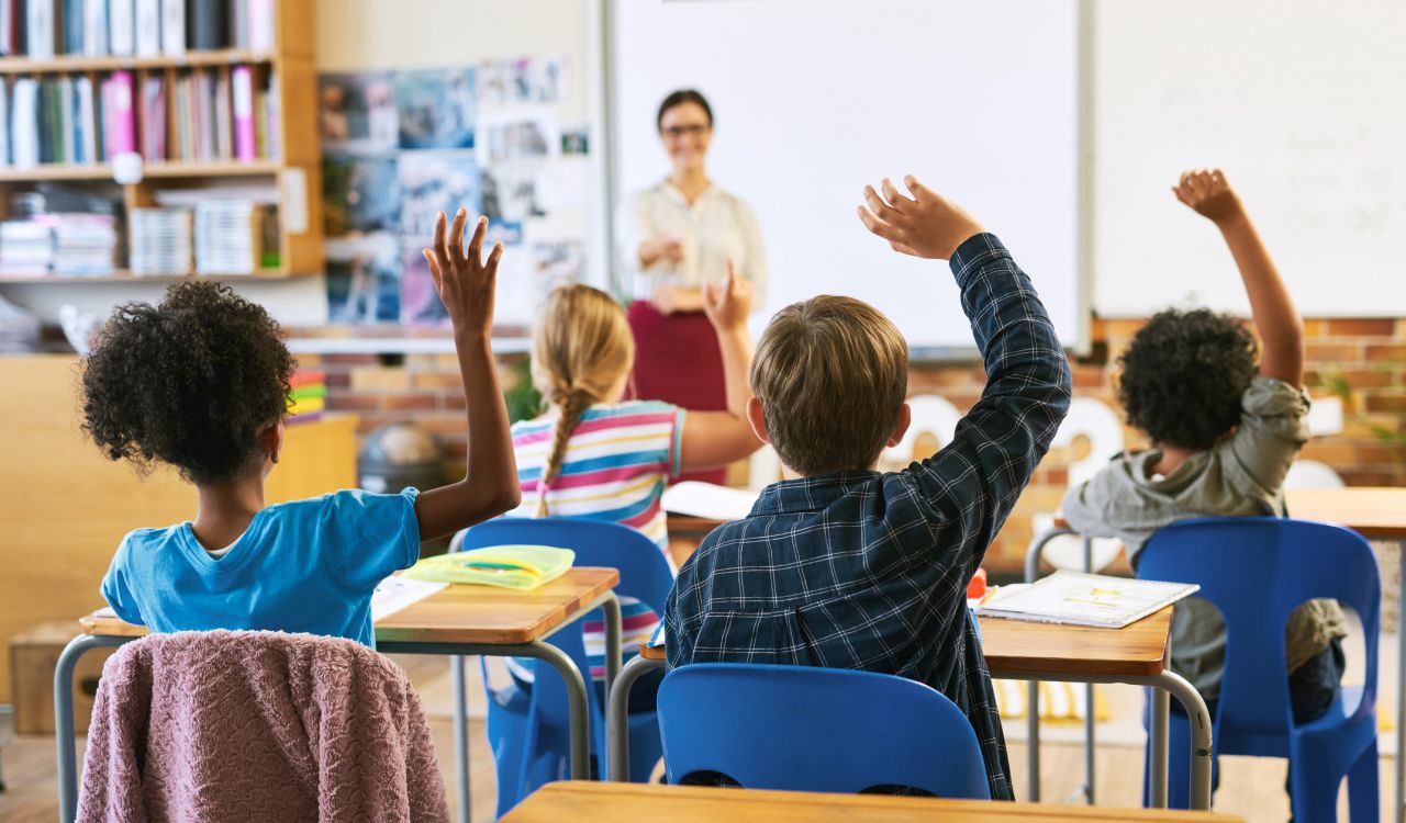 Children, seen from behind, raise their hands while sitting at desks in a classroom.