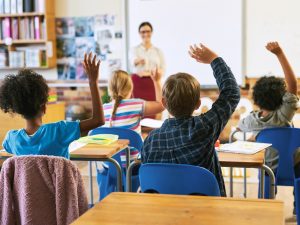 Children, seen from behind, raise their hands while sitting at desks in a classroom.