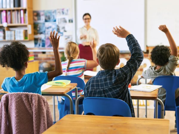 Children, seen from behind, raise their hands while sitting at desks in a classroom.