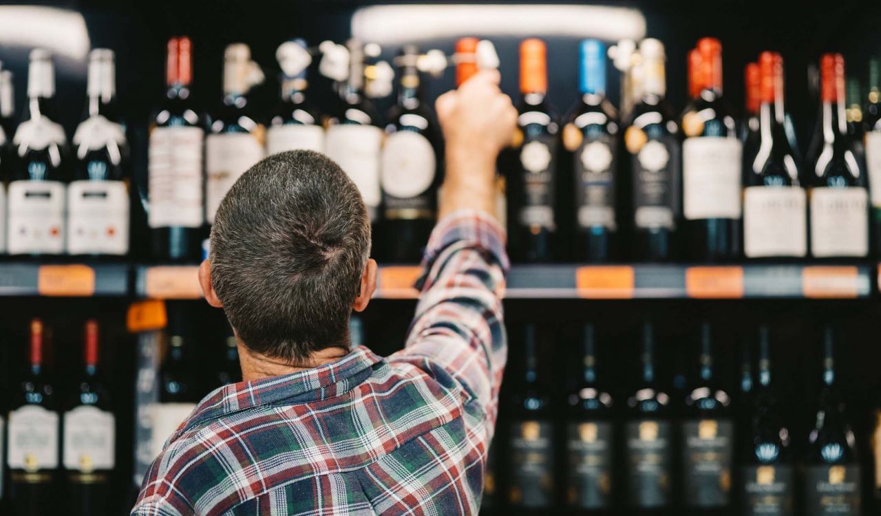 A person, seen from behind, reaches up to take a wine bottle off a shelf in a store.