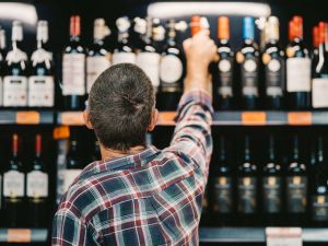 A person, seen from behind, reaches up to take a wine bottle off a shelf in a store.