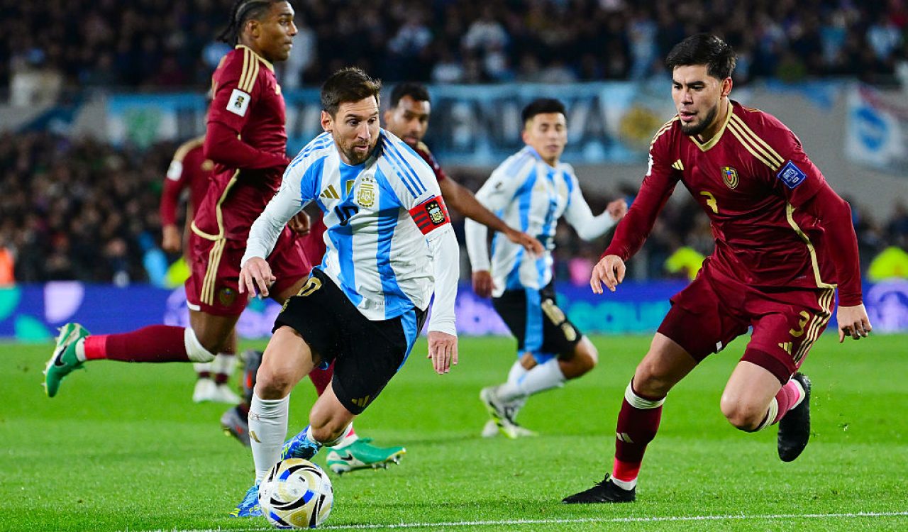 Soccer players from two teams on a green soccer pitch, one controlling the ball as others attempt to intervene.