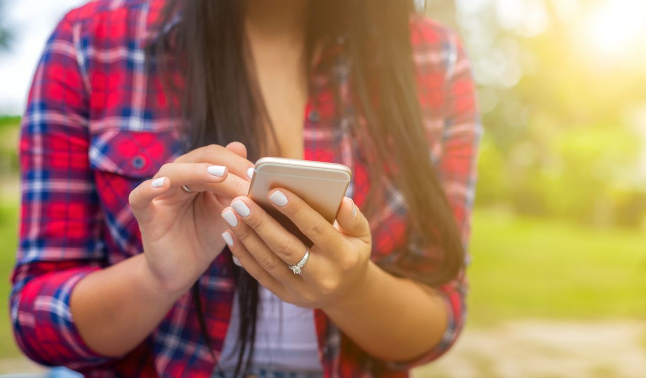 Close-up of hands holding and tapping a smartphone, with a red plaid shirt visible and a soft-focus outdoor background.