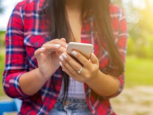 Close-up of hands holding and tapping a smartphone, with a red plaid shirt visible and a soft-focus outdoor background.