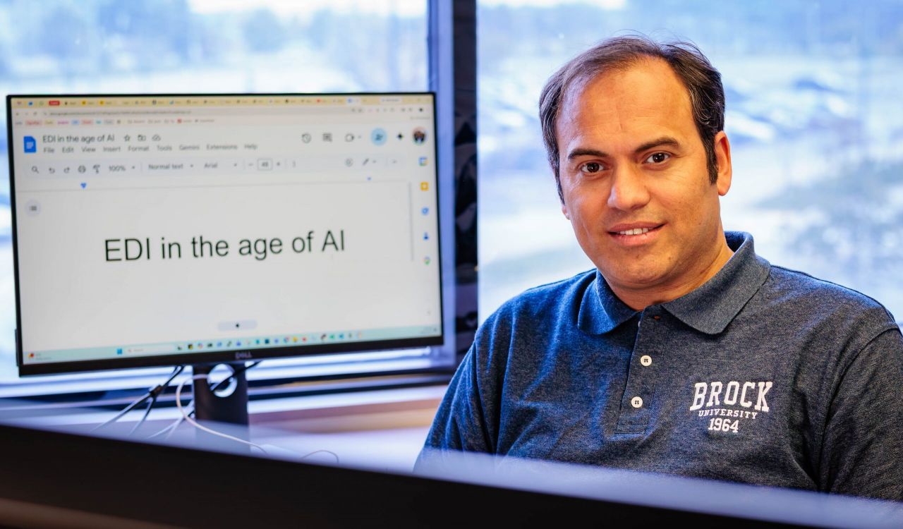 A man wearing a Brock University polo sits at a desk beside a computer monitor displaying the words ‘EDI in the age of AI.’