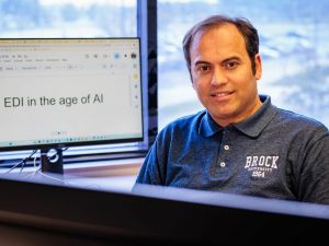 A man wearing a Brock University polo sits at a desk beside a computer monitor displaying the words ‘EDI in the age of AI.’