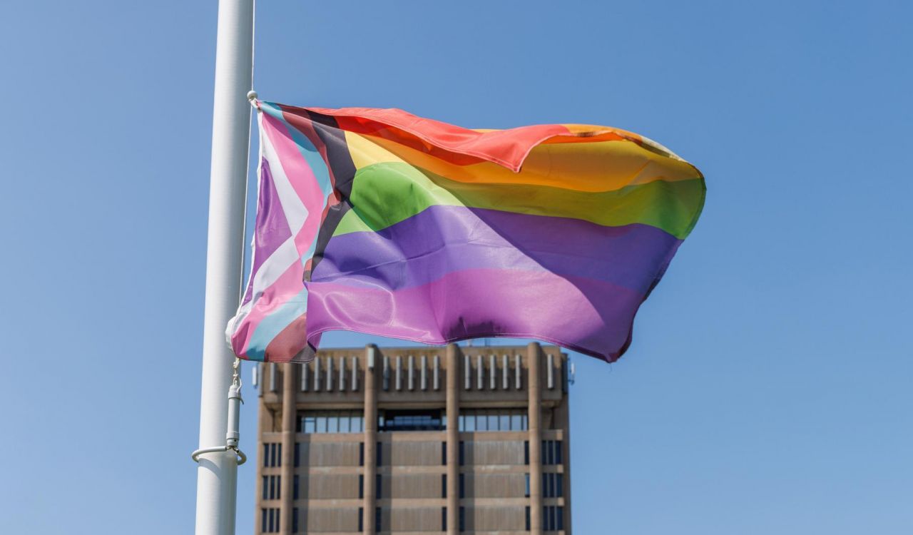 A colourful Pride flag flies in front of a concrete building with a blue sky in the background.