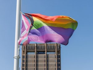 A colourful Pride flag flies in front of a concrete building with a blue sky in the background.
