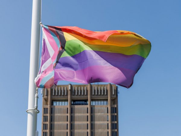A colourful Pride flag flies in front of a concrete building with a blue sky in the background.